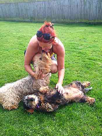 Jane on the grass with two dogs, one nose-to-nose with her and one having a tummy rub